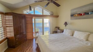 A bedroom with a large bed, a ceiling fan, a balcony with a view, and a wooden wardrobe.