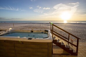 Un jacuzzi en una terraza de madera con vista a una playa y un atardecer de fondo.