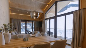 En modern inredning med ett matbord i trä, bekväma sittplatser, stora fönster med gardiner och utsikt över ett snötäckt landskap.