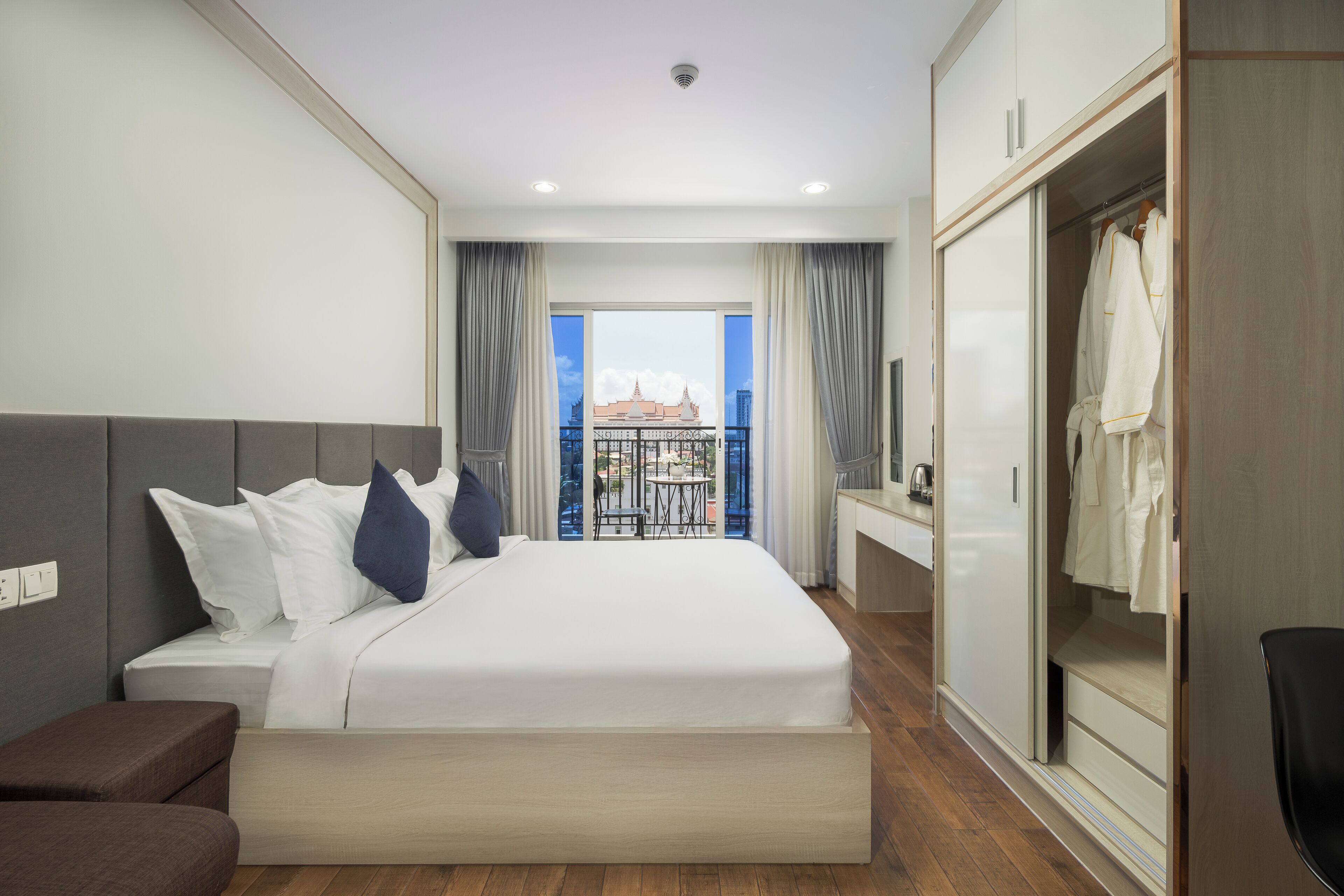 A hotel room with a large bed, a wardrobe, a seating area, and a balcony with a view of buildings.