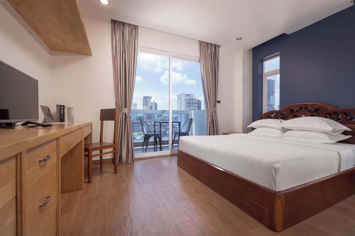 A hotel room with a wooden bed, a desk with a computer, a chair, and a balcony with a city view.