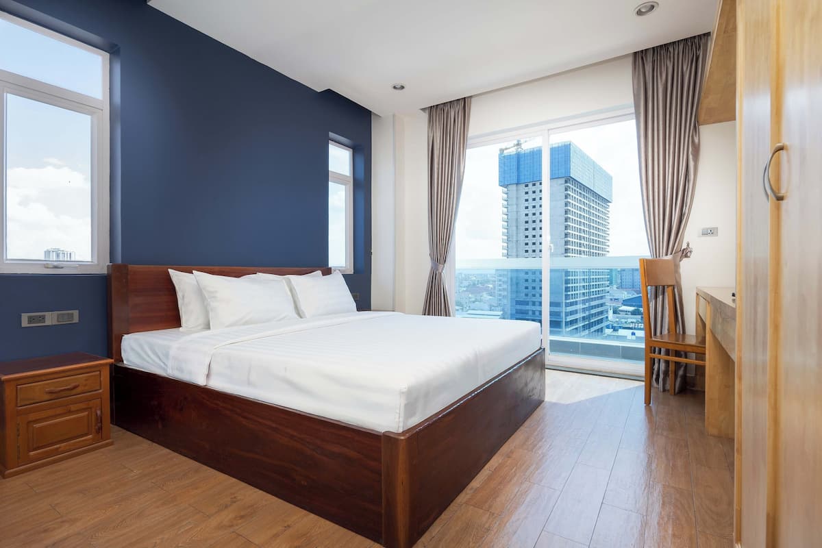 A modern hotel room with a large bed, wooden nightstand, and a view of a cityscape through a large window.