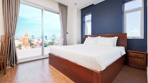 A hotel room with a large bed, wooden floor, a nightstand, a chair, and a view of a cityscape through large windows.