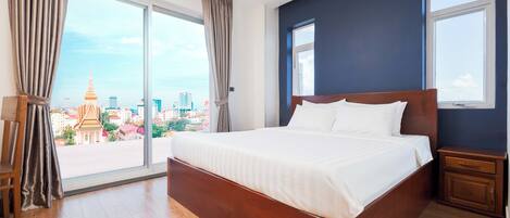 A hotel room with a large bed, wooden floor, a nightstand, a chair, and a view of a cityscape through large windows.