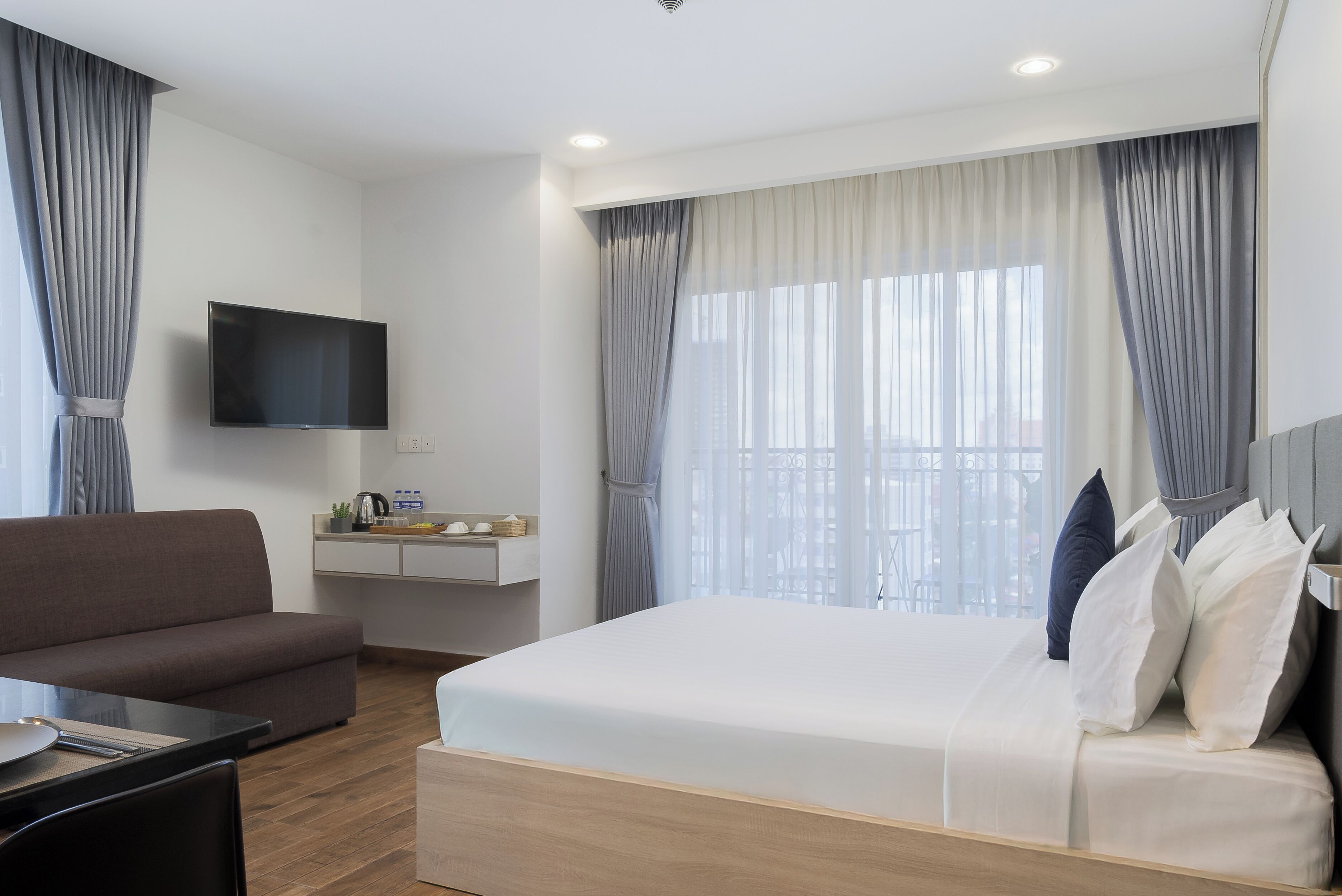 Quarto de hotel com cama, sofá, televisão fixada na parede, uma pequena mesa com chaleira e uma janela com cortinas.