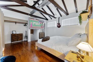 Une chambre avec un lit en bois, une table de chevet avec une lampe, une guitare, un téléviseur et une fenêtre avec des stores.