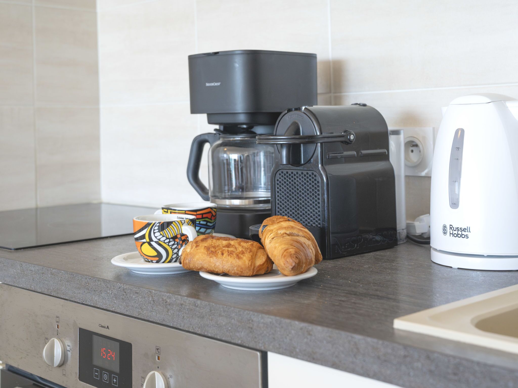 A kitchen with a coffee maker, a kettle, and croissants on the counter.