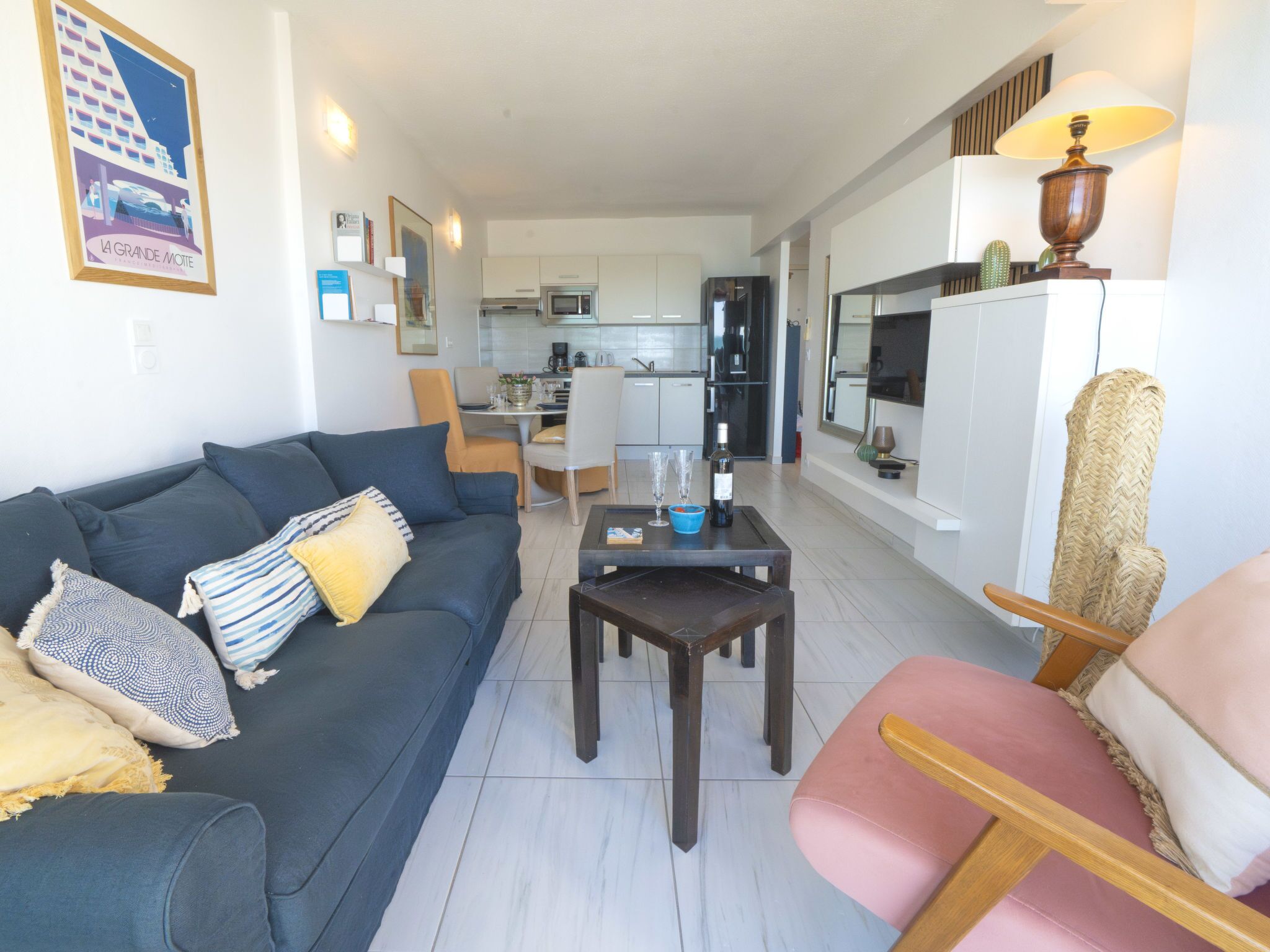 En moderne stue med en mørkeblå sofa, en lyserød lænestol og et spiseområde med bord og stole. Et køkken er synligt i baggrunden med hvide skabe og et køleskab.