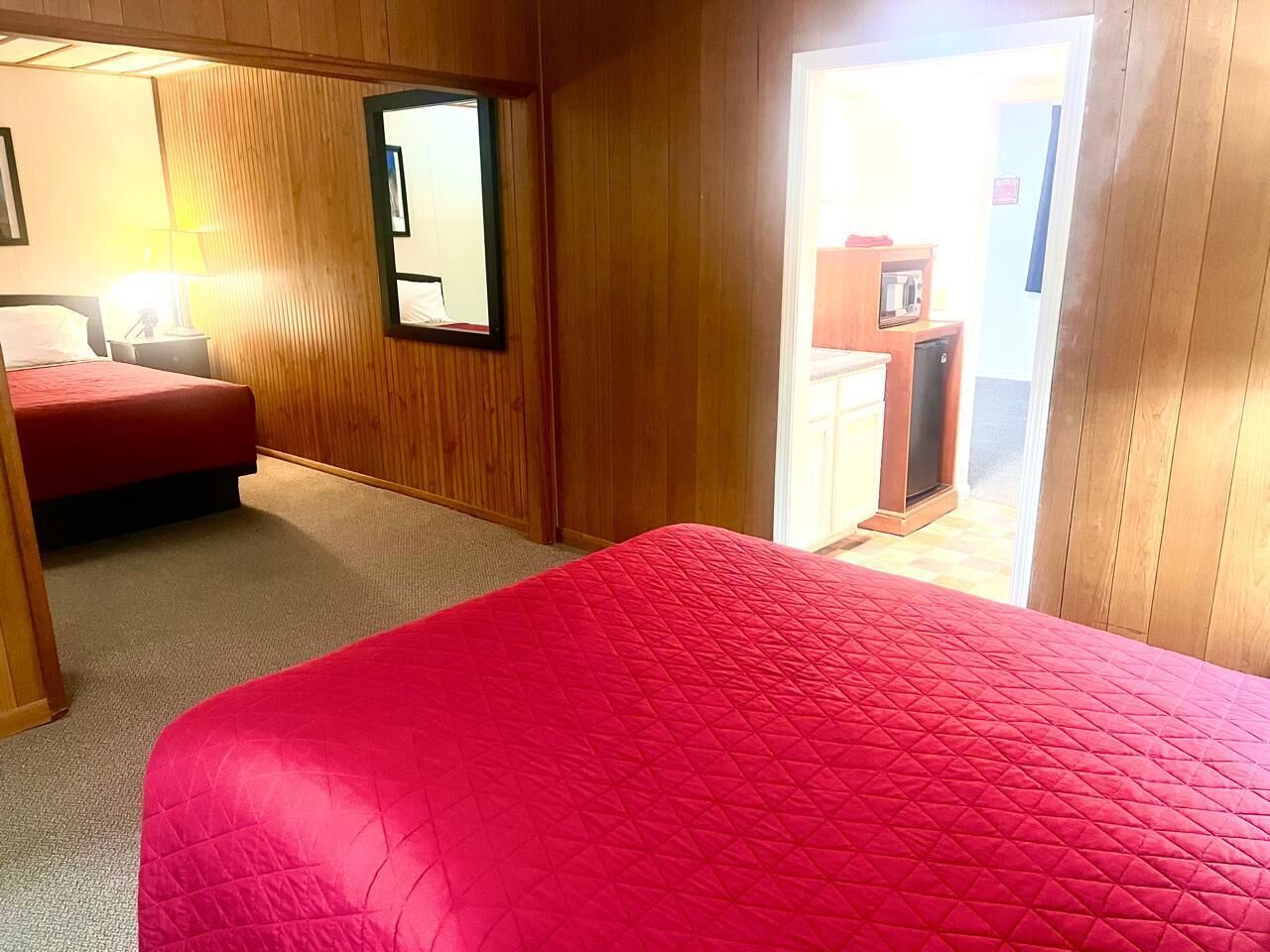 A hotel room with a large bed, a smaller bed, a wooden wardrobe, a mirror, and a kitchenette area.