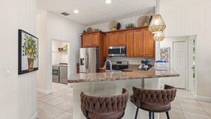 A kitchen with wooden cabinets, stainless steel appliances, granite countertops, and two upholstered bar stools. - Premium Modern Pool Home- 1 Mile to Downtown & 3 Miles to Beaches (Jensen Beach)