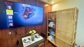 Habitación con televisión mostrando un perfil de Netflix, un icono de Airbnb y un cartel de Cazador Kraven. Hay un estante de madera con toallas y una mesita con un jarrón de flores.