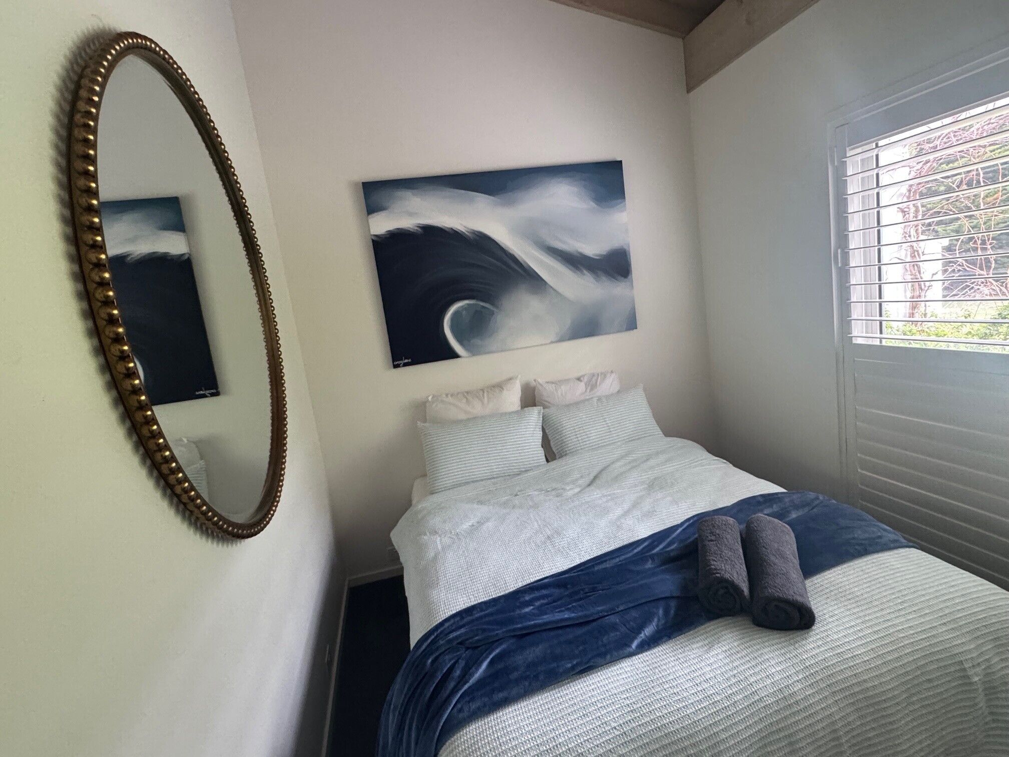Une chambre à coucher avec un lit, un grand miroir, un tableau et une fenêtre avec des stores.
