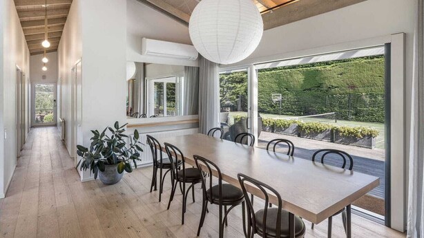 Un espace repas moderne avec une longue table et des chaises, de grandes fenêtres donnant sur un jardin aménagé, et un luminaire suspendu.