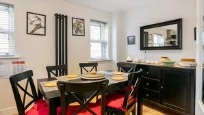 Een eetruimte met een tafel voor vier personen, zwarte stoelen met rode kussens, een zwarte kast met spiegel en twee ingelijste kunstwerken aan de muur.