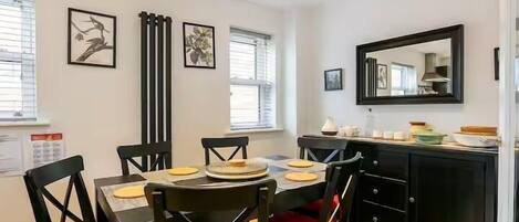 A dining area with a table set for four, black chairs with red cushions, a black cabinet with a mirror, and two framed artworks on the wall.