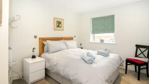 Een slaapkamer met een bed, nachtkastje, stoel, raam met uitzicht en een foto aan de muur.