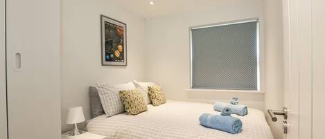 A bedroom with a bed, pillows, a nightstand, a lamp, a poster on the wall, and a window with blinds.
