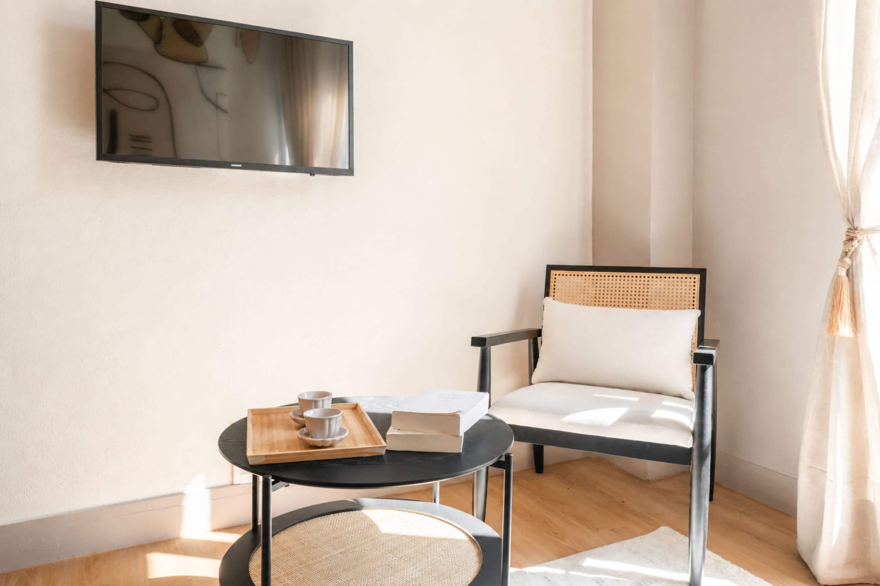 Een moderne kamer met een flatscreen televisie aan de muur, een ronde tafel met een dienblad met kopjes en een boek, en een stoel met een geweven rugleuning.
