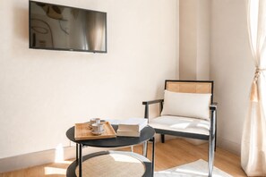 Une chambre moderne avec un téléviseur à écran plat fixé au mur, une table ronde sur laquelle se trouve un plateau avec des tasses et un livre, et une chaise dotée d’un dossier en osier.
