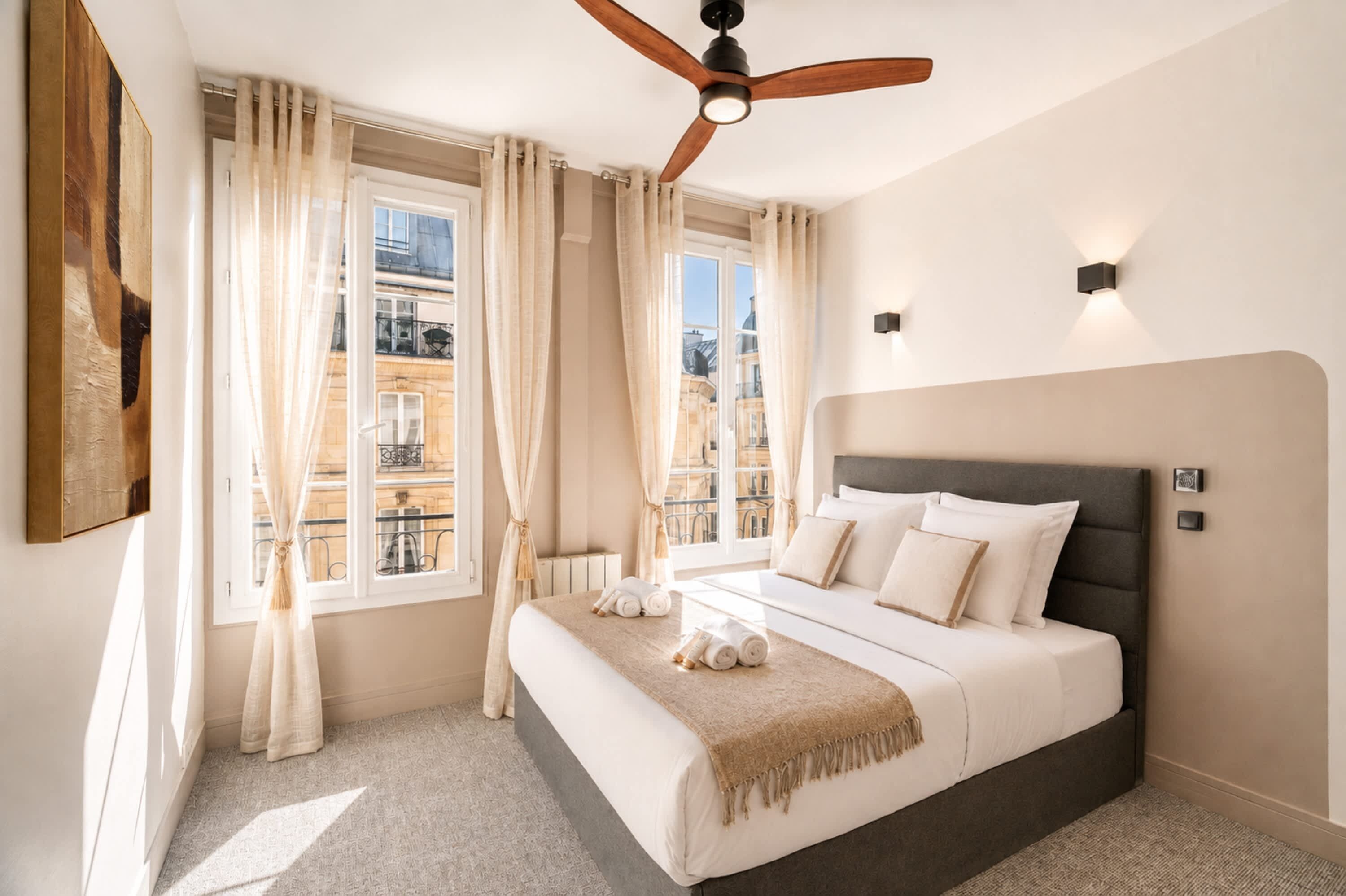 Een hotelkamer met een bed, gordijnen, een plafondventilator en uitzicht op gebouwen.