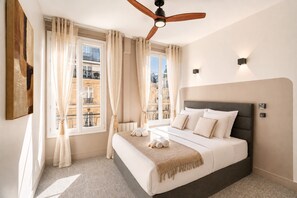 Une chambre d’hôtel avec un lit, des rideaux, un ventilateur de plafond et une vue sur les bâtiments environnants.