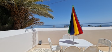 Una terraza con una mesa y sillas blancas, un paraguas colorido y vistas al mar con barcos.