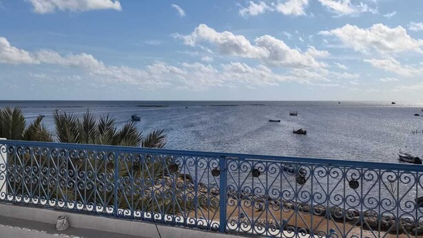 A coastal view with a metal railing, palm trees, and boats on the water. - Beit El Ezz Kerkennah  (Kerkennah Islands)
