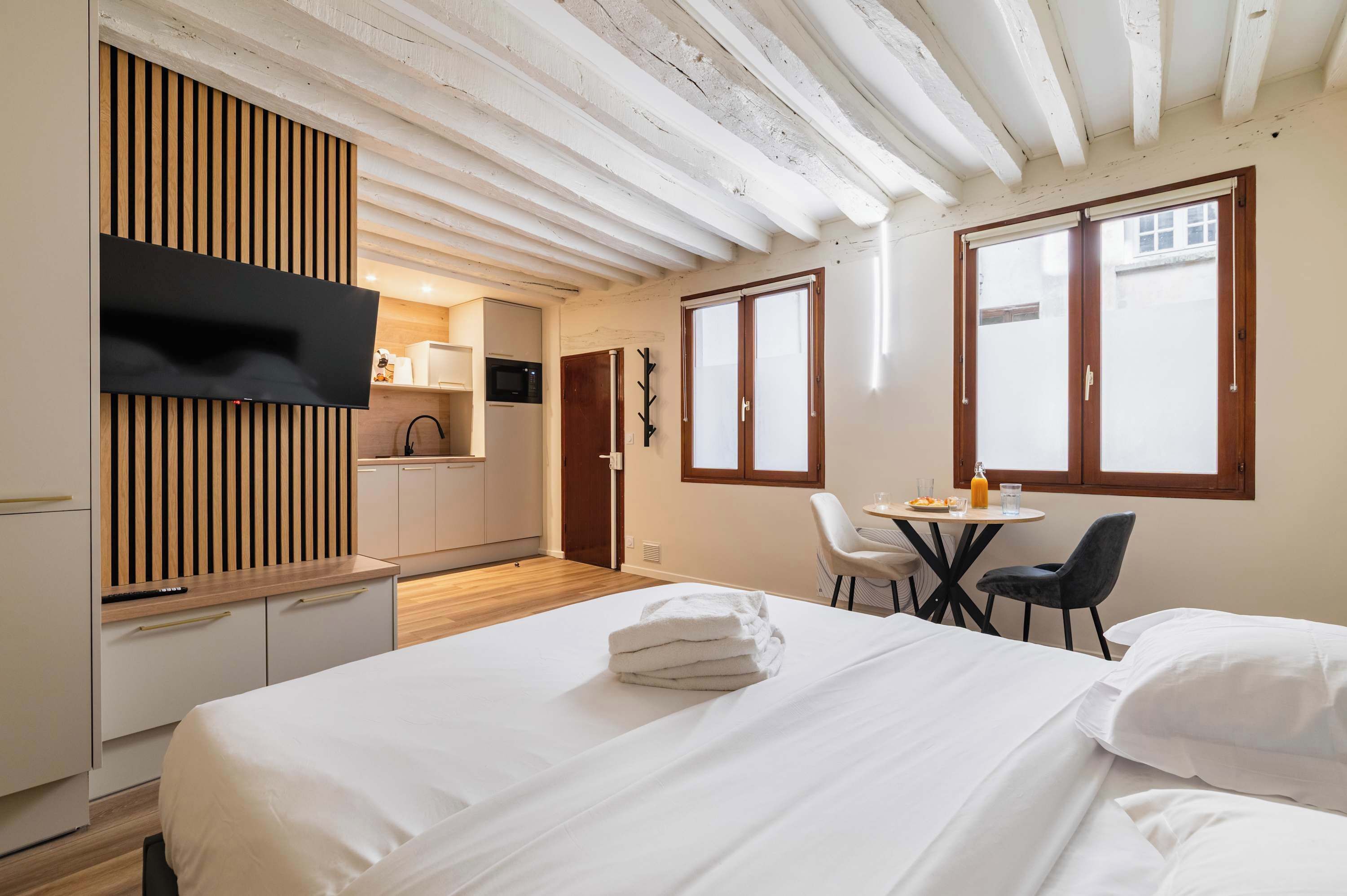 A hotel room with a bed, a TV mounted on a wooden panel, a kitchenette, and a dining area with a table and chairs.