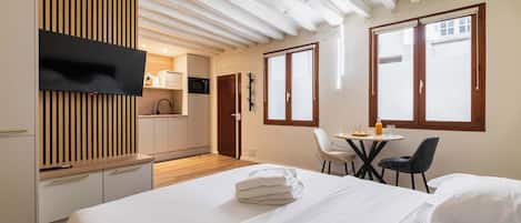 A hotel room with a bed, a TV mounted on a wooden panel, a kitchenette, and a dining area with a table and chairs.