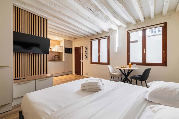 A hotel room with a bed, a TV mounted on a wooden panel, a kitchenette, and a dining area with a table and chairs.