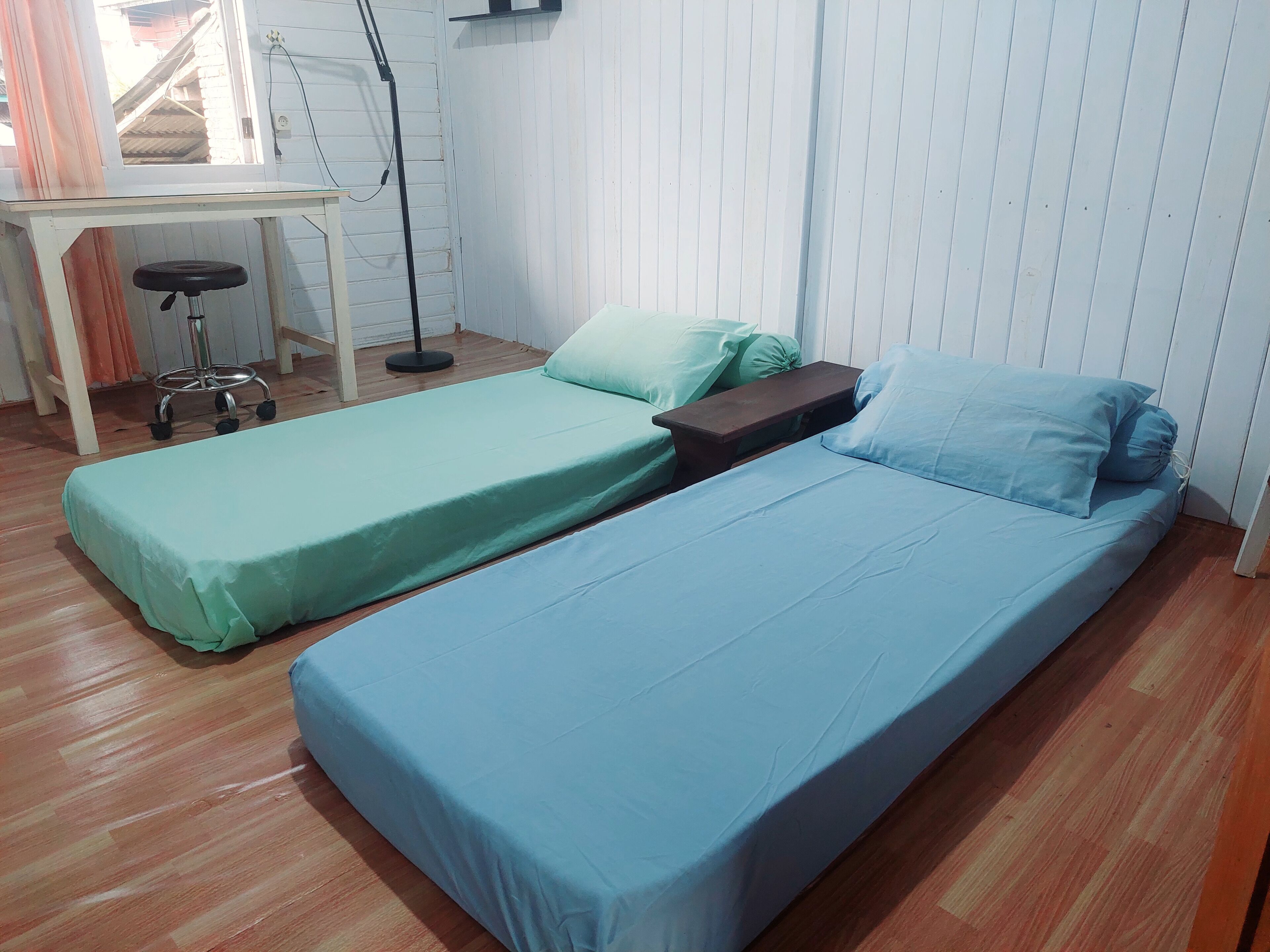 A room with two single beds, a small desk with a chair, and a standing lamp.
