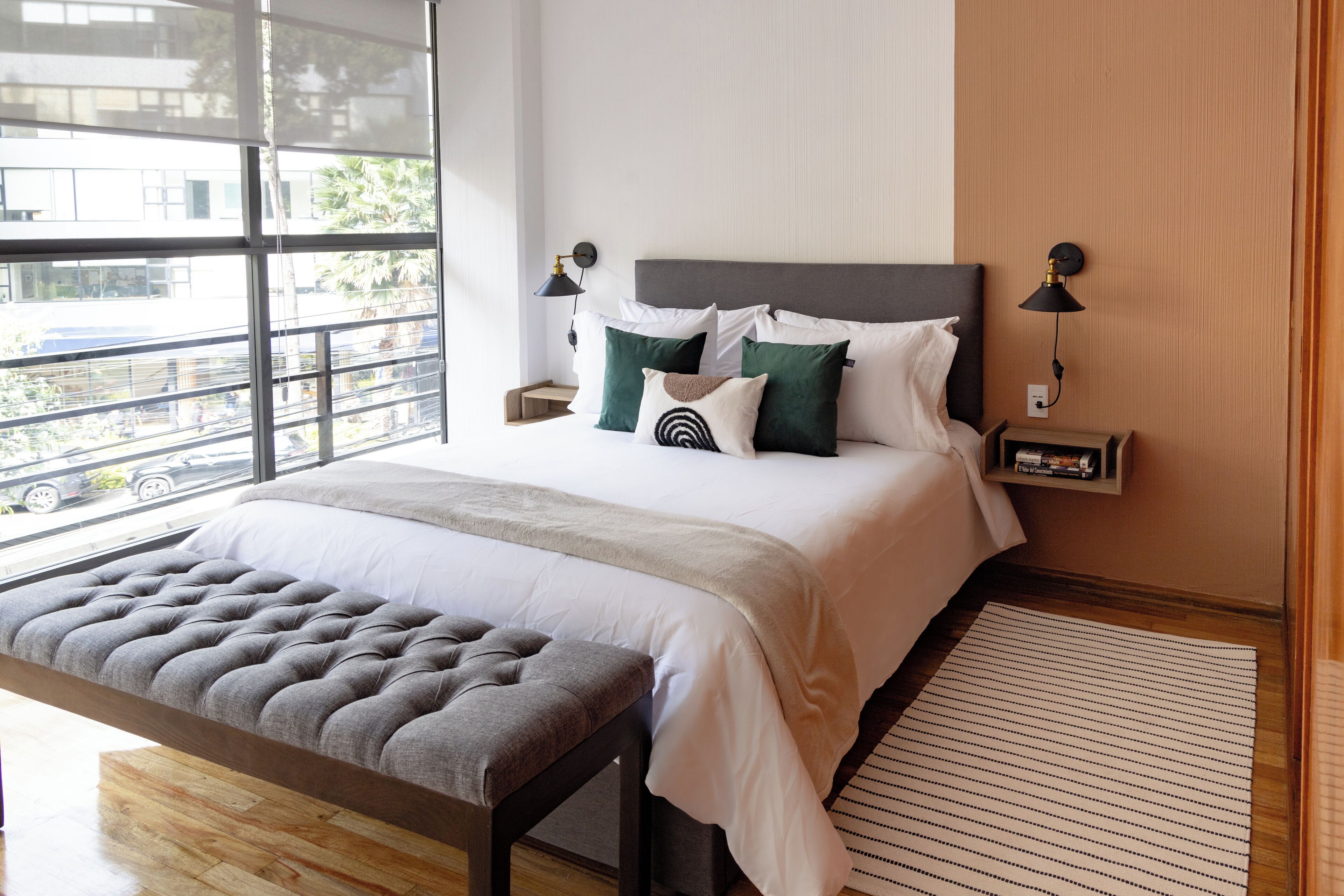 Habitació d'hotel moderna amb un llit gran, una banca i vista al carrer a través de finestres grans.