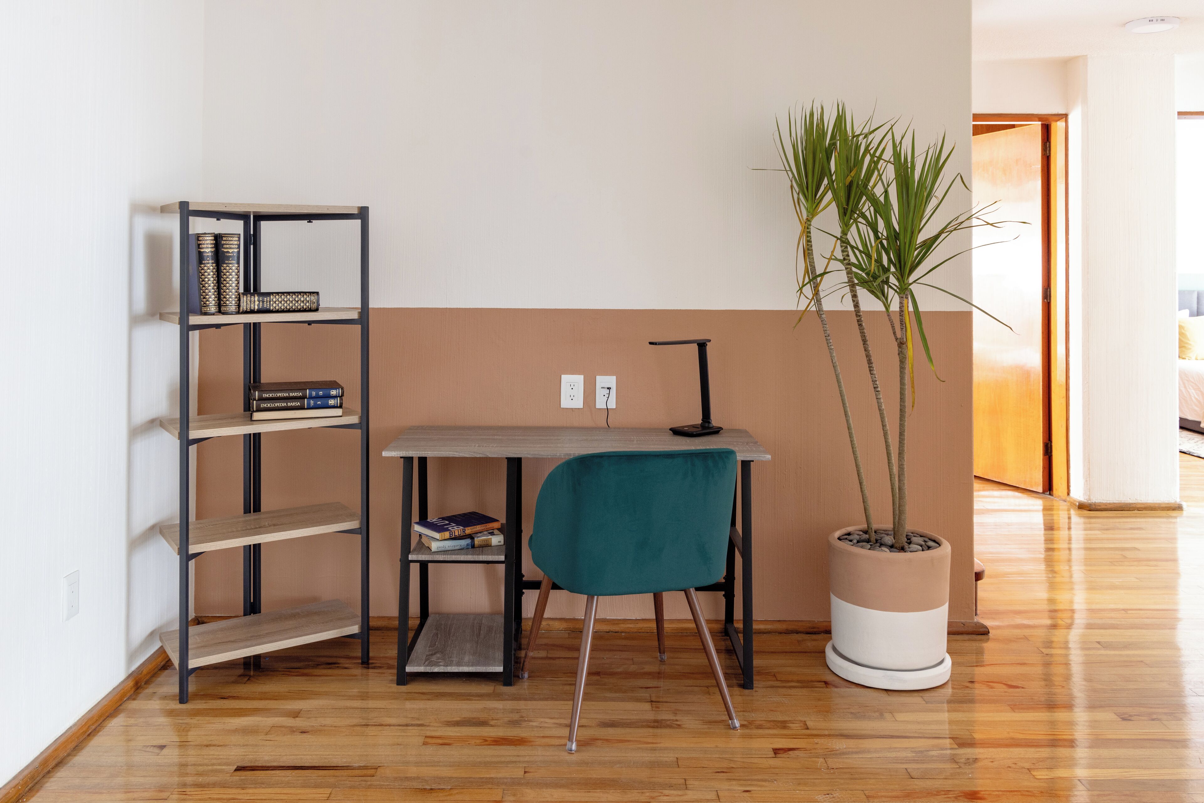 Una habitación con piso de madera, un escritorio con una silla color turquesa, una estantería y una planta en maceta.