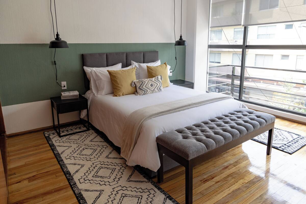 Un dormitorio ordenado con cama, mesitas de noche, un banco, un tapete estampado y un amplio ventanal con vistas a la ciudad.