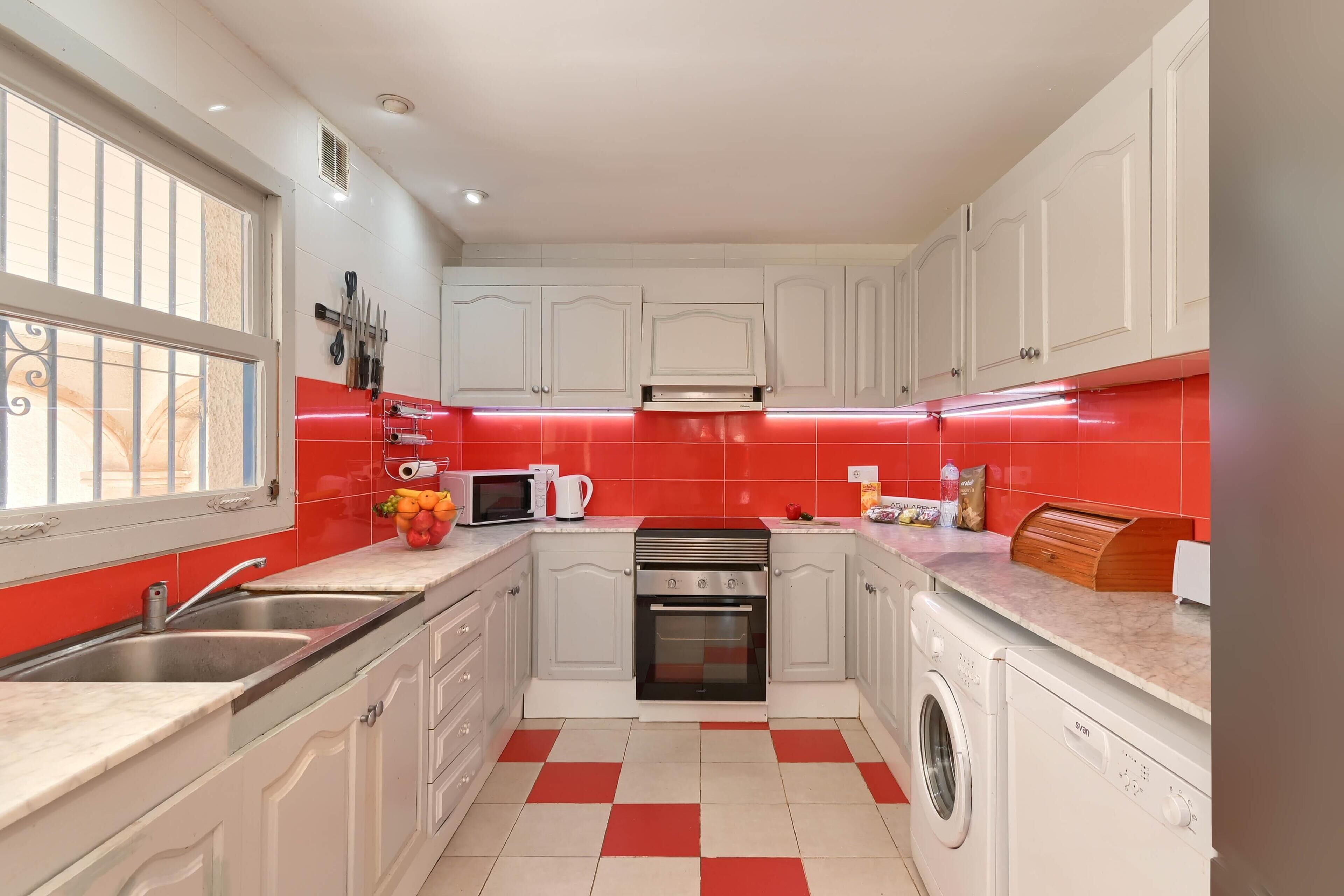 Una cocina con armarios blancos, salpicadero rojo, microondas, lavaplatos, estufa y lavadora.