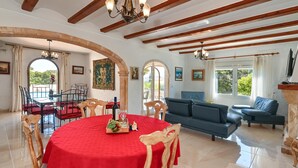 A spacious living area with a round table covered in a red tablecloth, wooden chairs, a blue sofa, and a chandelier. - Ker Blanc 8 pax - Javea, Costa Blanca (Javea)