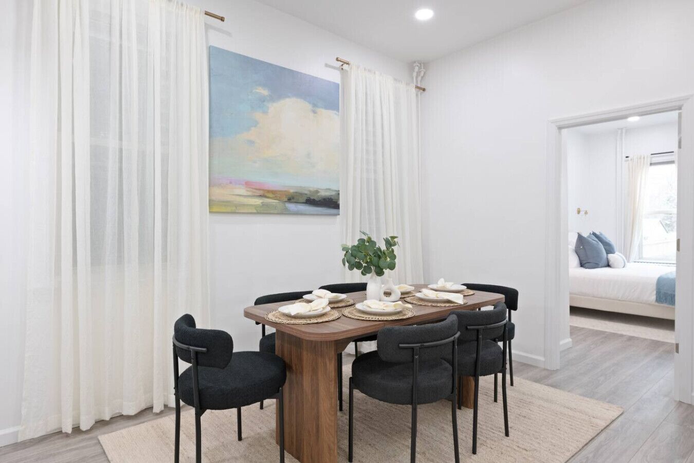 Een eetruimte met een houten tafel, zwarte stoelen en een wit kleed. Aan de muur hangt een schilderij en er is een raam met gordijnen.