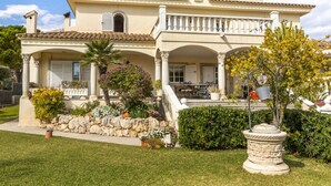 A Mediterranean-style villa with a tiled roof, a balcony with white balustrades, and a stone wall with various plants.