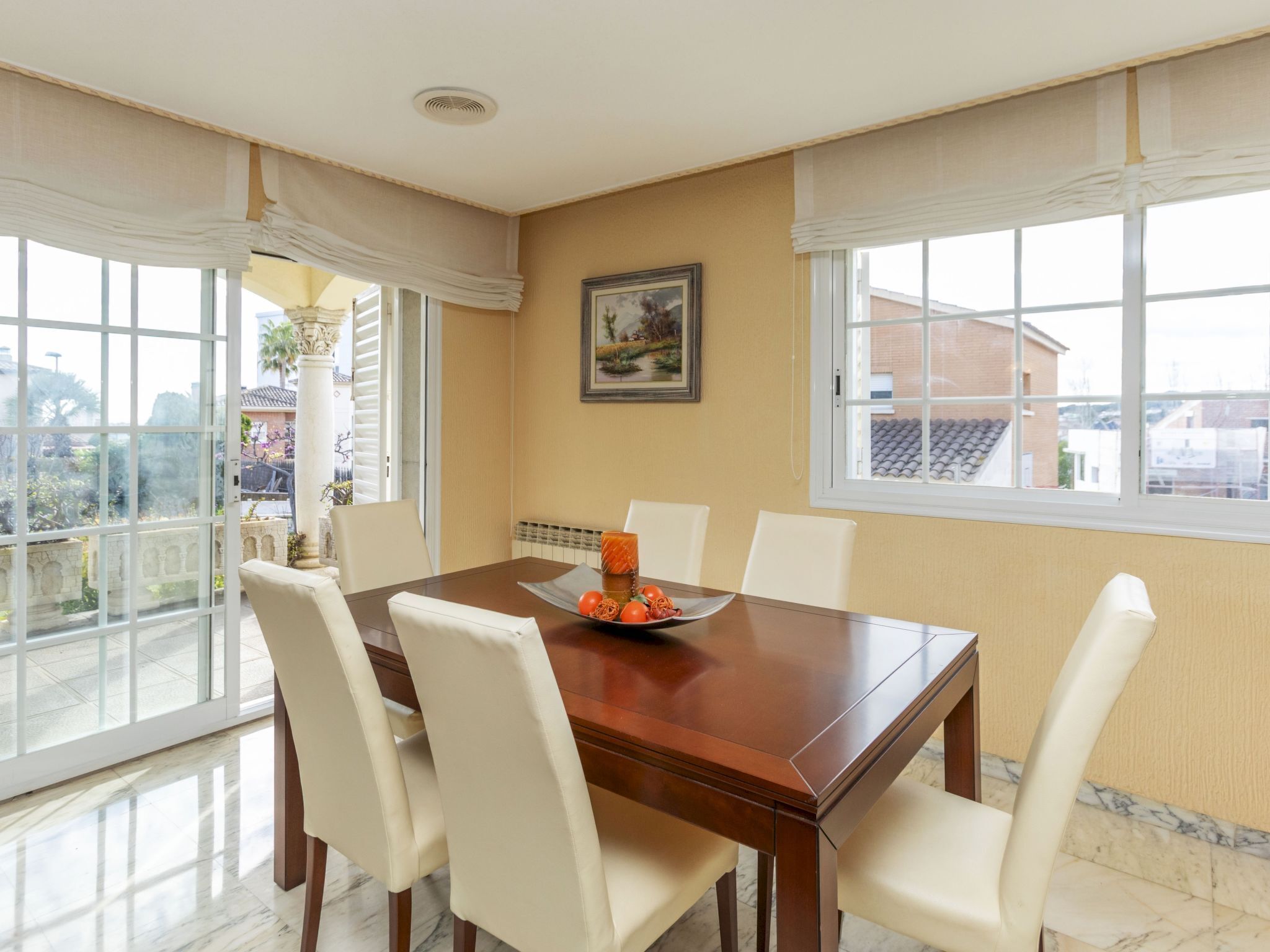 A dining room with a wooden table, white chairs, a painting on the wall, and large windows with a view of buildings and greenery.