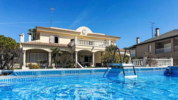 A swimming pool with clear blue water, a white fence, and a large two-story building with a balcony in the background.