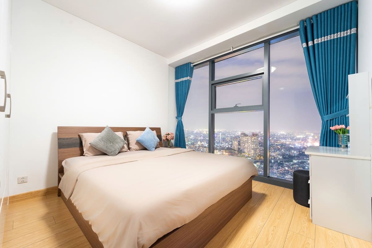 A bedroom with a large bed, a desk, and a view of a cityscape through a large window.