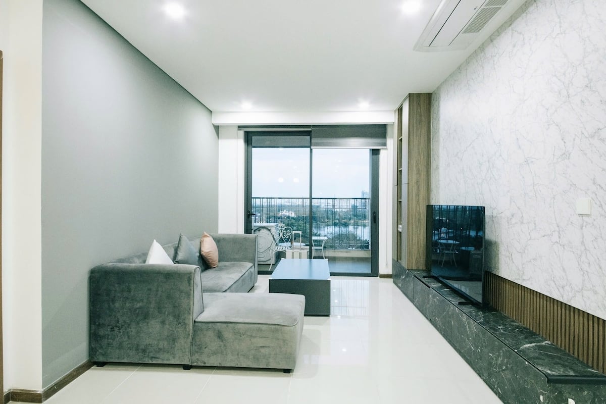 A modern living room with a grey sofa set, a flat-screen TV, and a balcony with a city view.