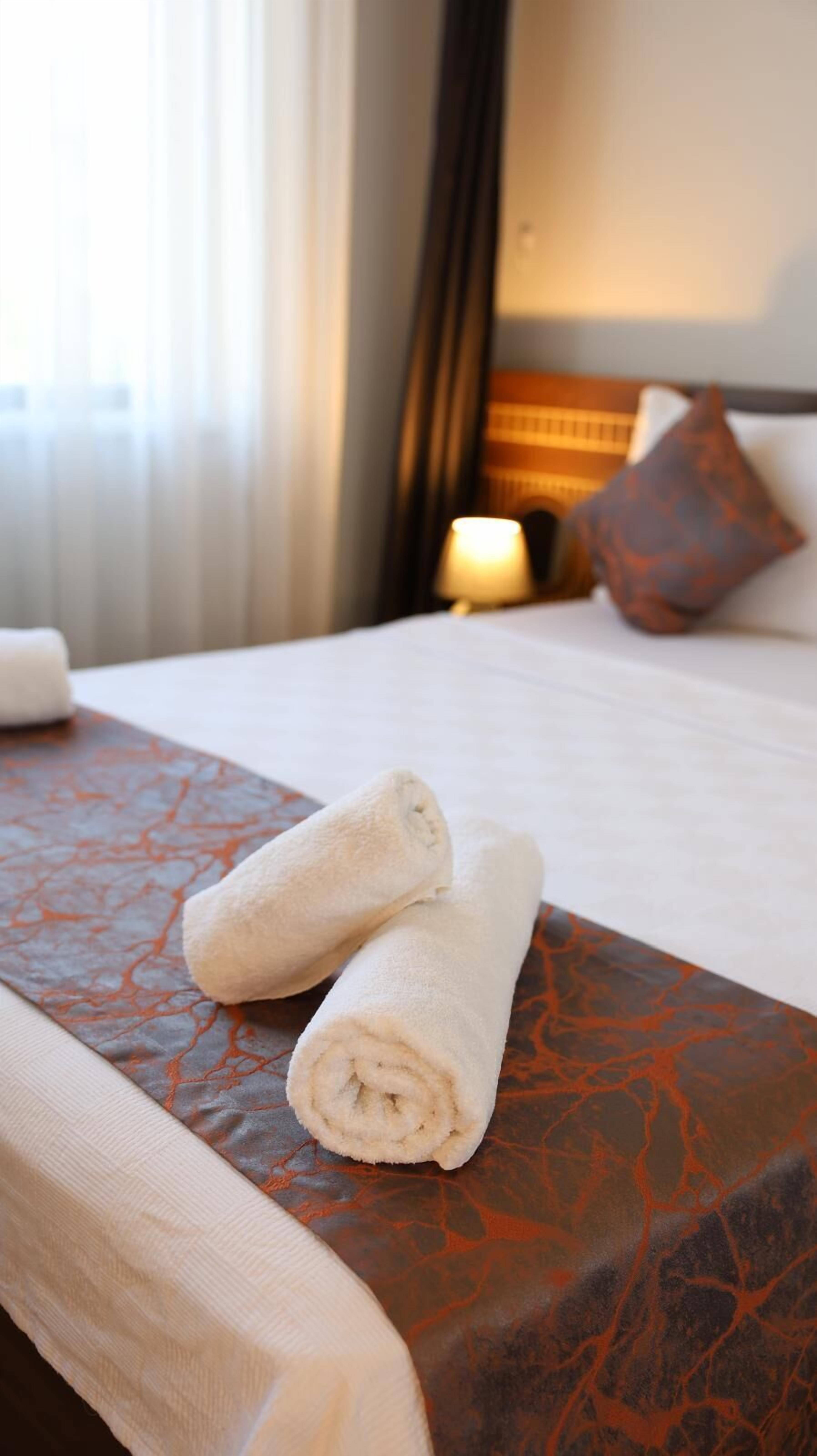 A neatly made bed with two rolled towels on a patterned bedspread, a bedside lamp, and a pillow in a hotel room.