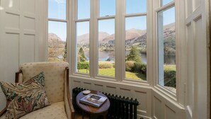 A room with a bay window overlooking a lake and hills. There is a chair with a cushion and a small table with a book. - Huntingstile South (Ambleside)