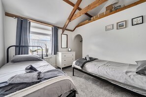 Een slaapkamer met twee bedden, een raam met gordijnen, een nachtkastje met een lamp, een dressoir en lijstjes met foto’s aan de muur.