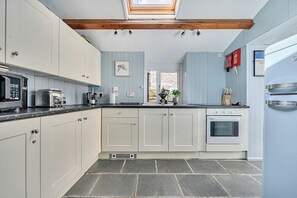 A kitchen with white cabinets, a black countertop, a built-in oven, and a window above the sink. - Mews Cottage (Tintagel)