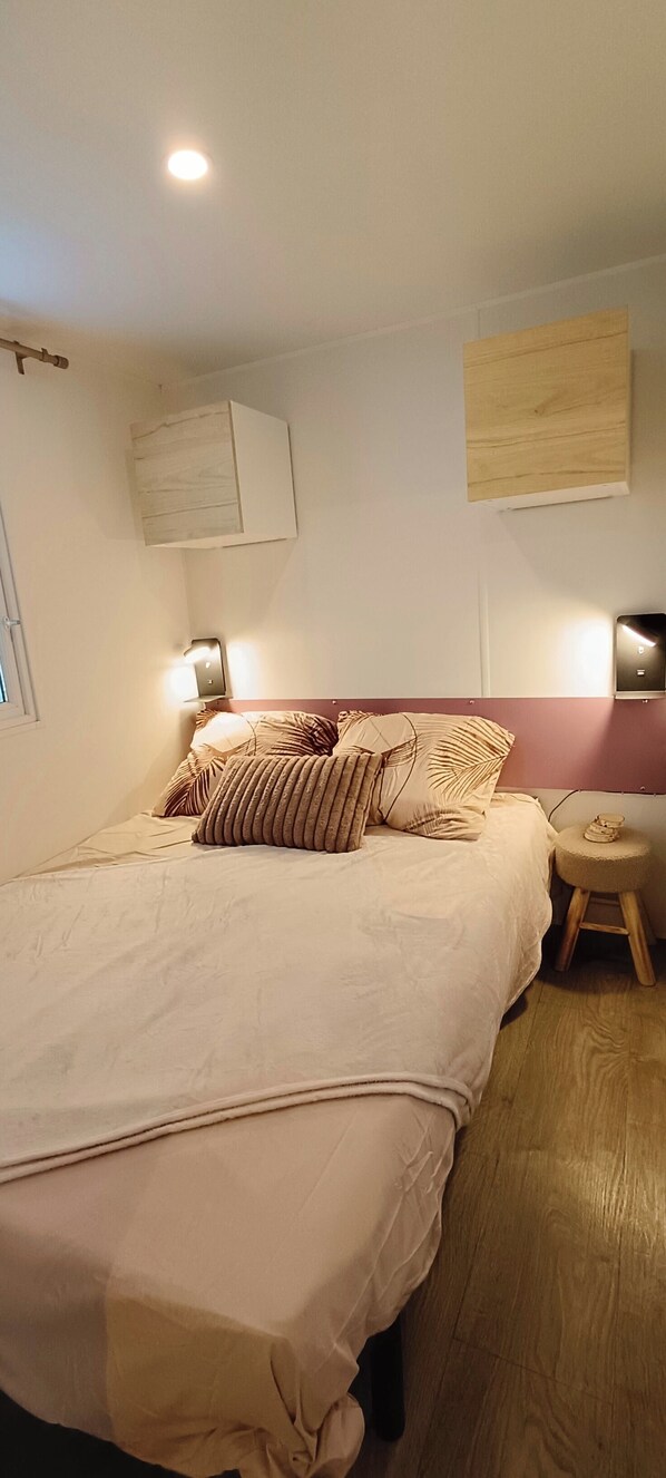 A neatly made bed with a beige comforter and striped pillows, a small wooden stool, and a wall-mounted light fixture.