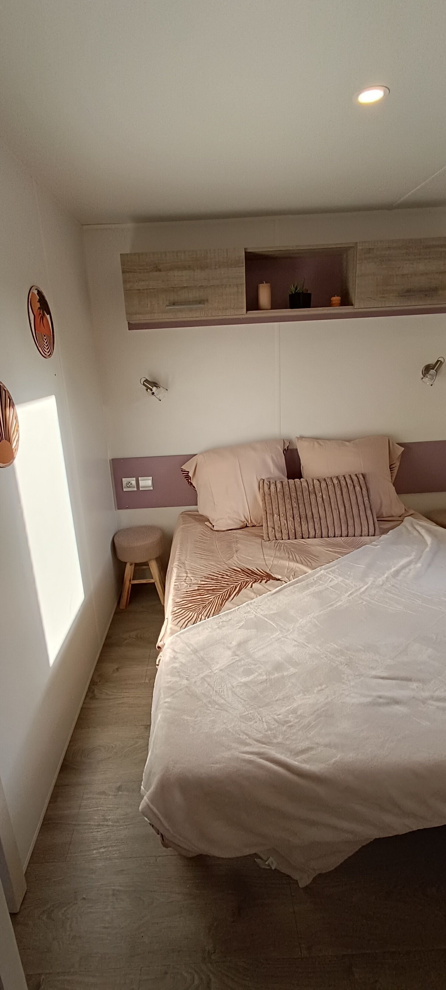 Et soveværelse med en seng, en lille skammel, en hylde over sengen og en lampe på væggen.