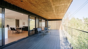 A modern outdoor balcony with a wooden deck, glass doors leading to an interior dining area, and a view of trees. - Tree Top Haven ITAS - Hosted by L'Abode (South Launceston)