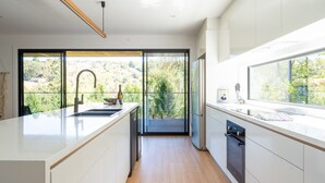 A modern kitchen with a large island, a built-in oven, and a view of a balcony with a scenic landscape. - Tree Top Haven ITAS - Hosted by L'Abode (South Launceston)